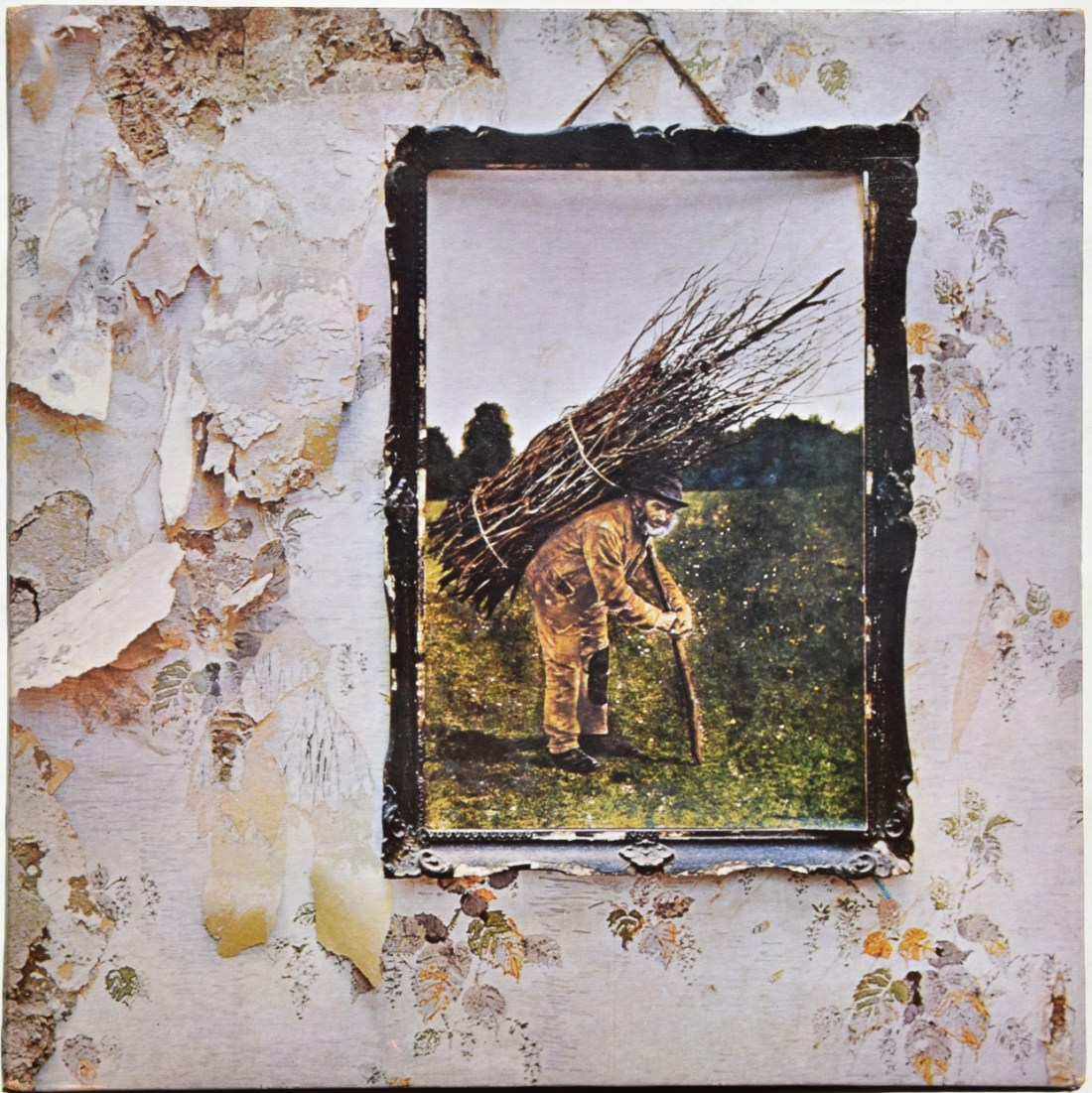 Una foto incorniciata attaccata a un muro malmesso, con la carta da parati tutta strappata. Nella foto è raffigurato un uomo abbastanza anziano piegato dal peso di una fascina che si regge a un bastone