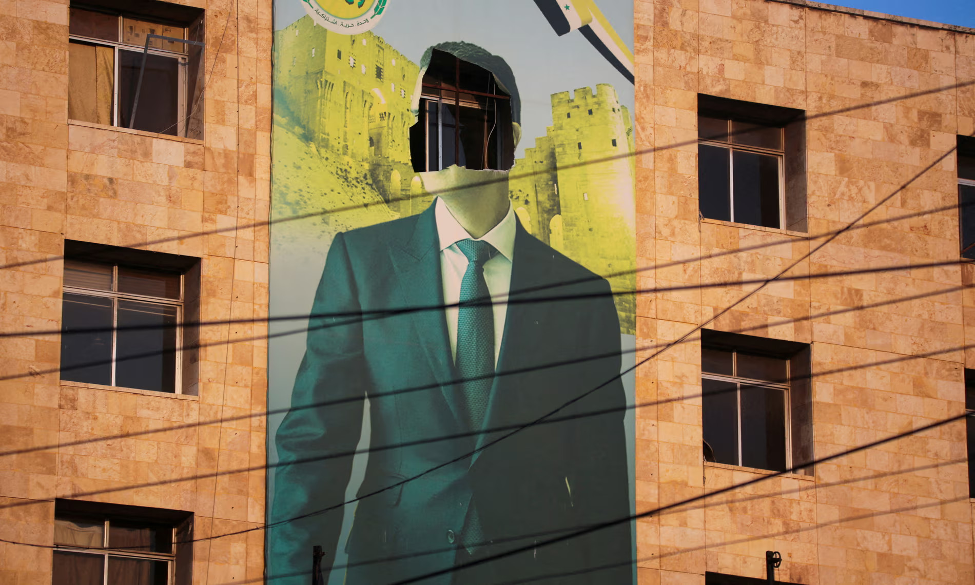 La foto di un poster gigante raffigurante Bashar al-Assad, senza faccia. Accanto al poster, a destra e sinistra, si vedono i muri rossicci e le finestre del palazzo sul quale il poster è affisso