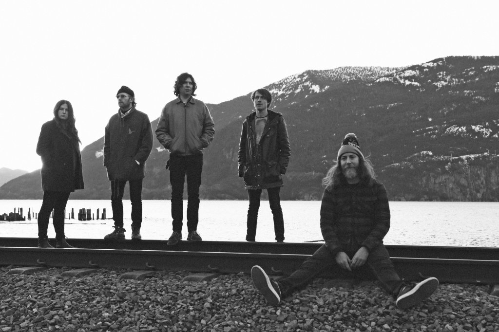 The Black Mountain are standing on railway tracks. One of them, on the right, is sitting on the track with his legs spread out and stretched out on the gravelly ground next to the track. Behind the band is what looks like a lake and a slightly snow-capped mountain.